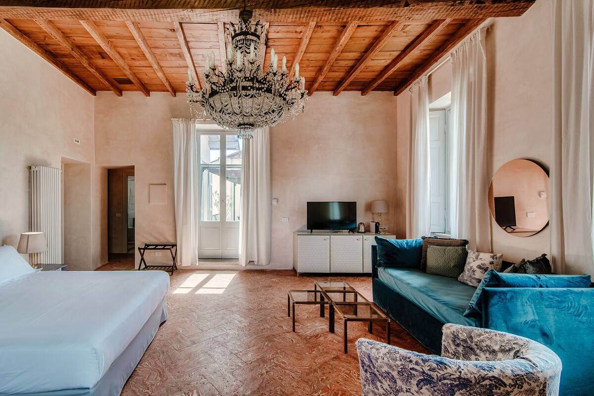A hotel room with a wooden ceiling, a chandelier, a bed, a sofa, a TV, and a window with curtains.