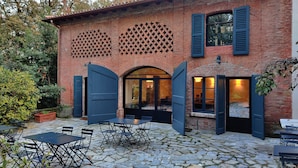 Un edificio rústico con fachada de ladrillo, amplias ventanas de arco y un patio empedrado con mesas al aire libre.