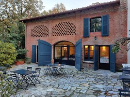 Un edificio rústico con fachada de ladrillo, amplias ventanas de arco y un patio empedrado con mesas al aire libre.