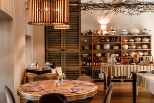 Un comedor con una mesa de madera dispuesta para dos personas, una estantería de madera con varios platos y botellas, y una lámpara colgante.