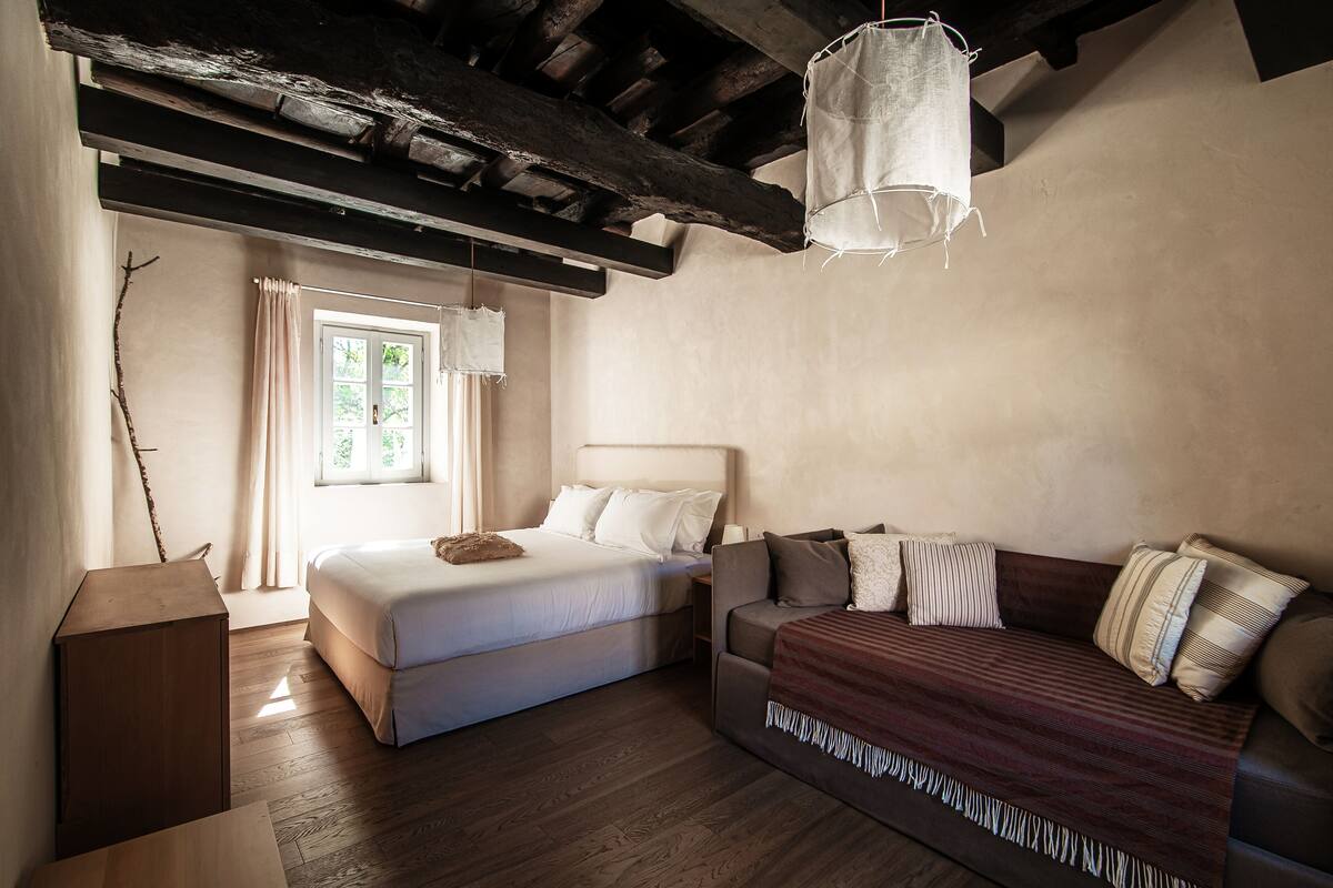 A bedroom with a bed, a sofa, a wooden floor, a window with curtains, and a hanging light fixture.