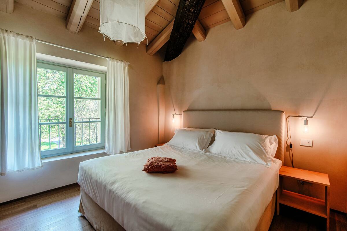 A bedroom with a large bed, a nightstand, a lamp, and a window with curtains.