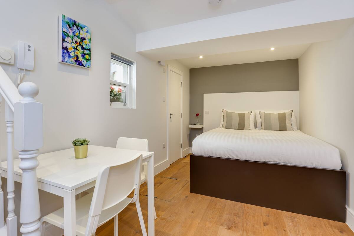 A bedroom with a bed, a bedside table, a chair, a small potted plant, a window, and a painting on the wall.