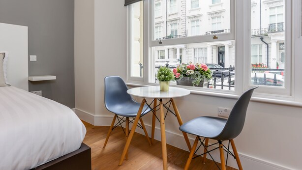 A modern hotel room with a large bed, two blue chairs, a small round table, and a window with a city view.