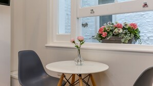 A small round table with a vase of pink flowers, a gray chair, and a window with a view of a building and flowers outside.