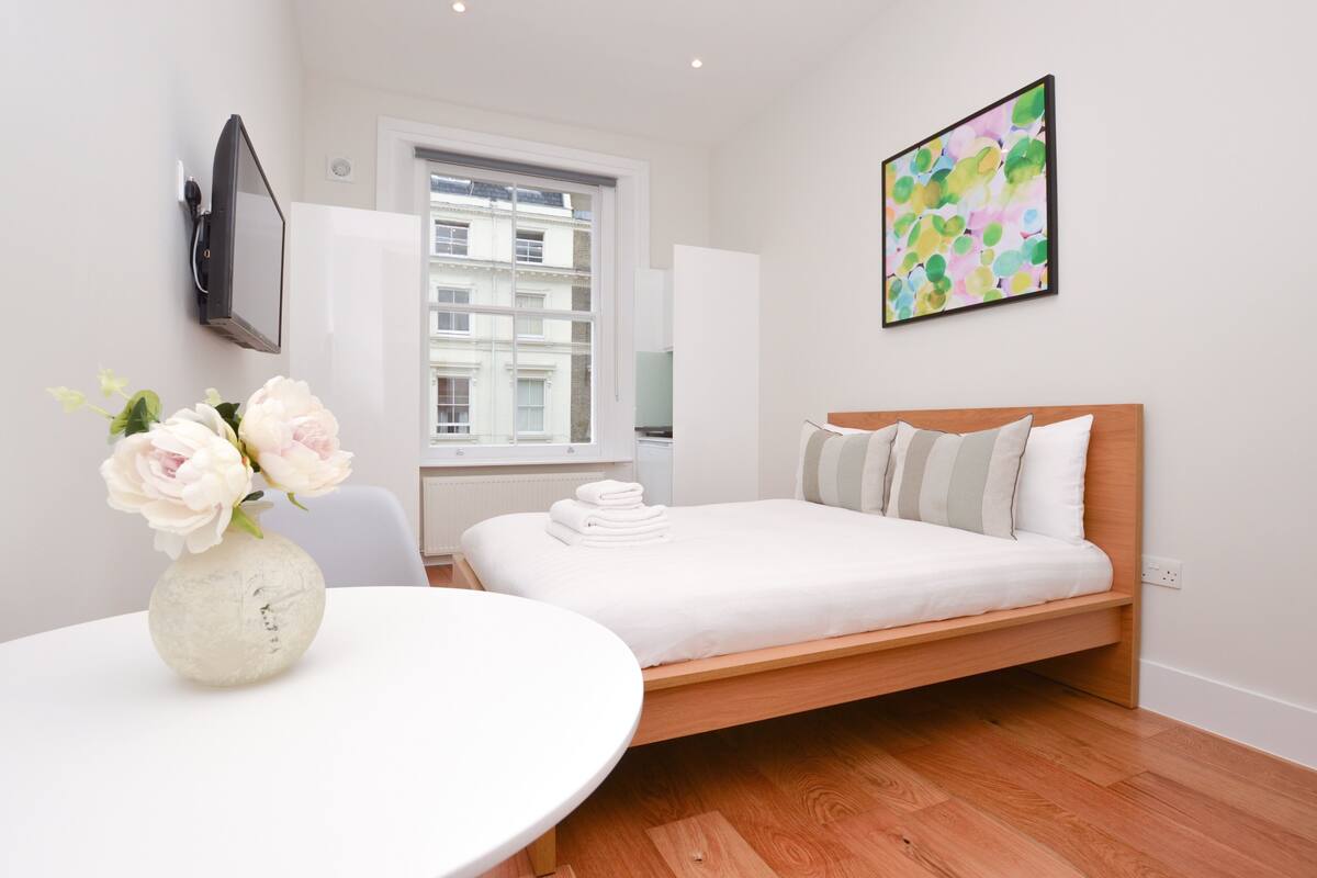 A modern bedroom with a bed, a round table with a vase of flowers, a window with a view of buildings, and a colorful abstract painting on the wall.