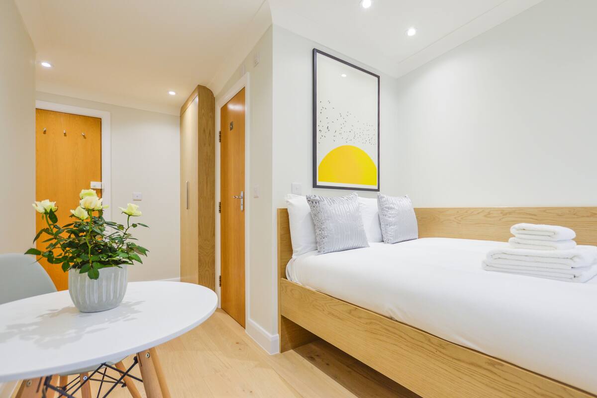 A modern hotel room with a wooden bed, a round table with a potted plant, and a large framed artwork on the wall.