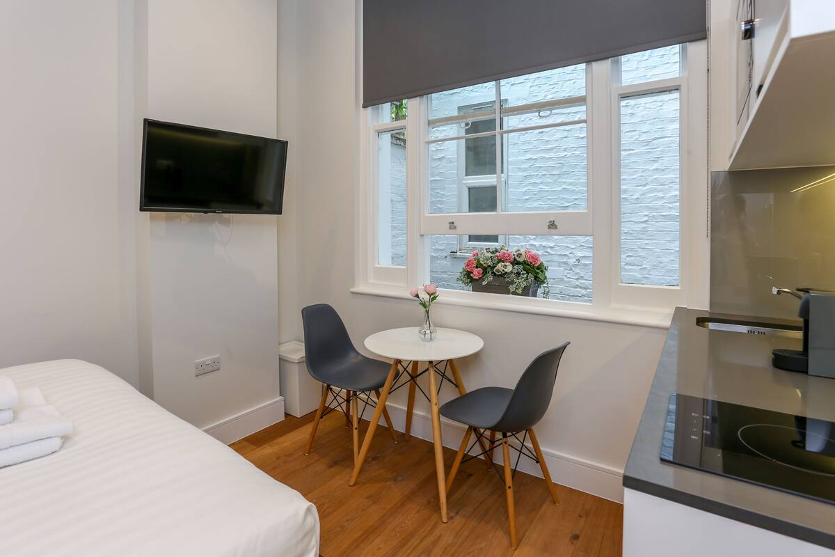 A modern room with a bed, a TV mounted on the wall, a small round table with two chairs, and a window with a view of a building.