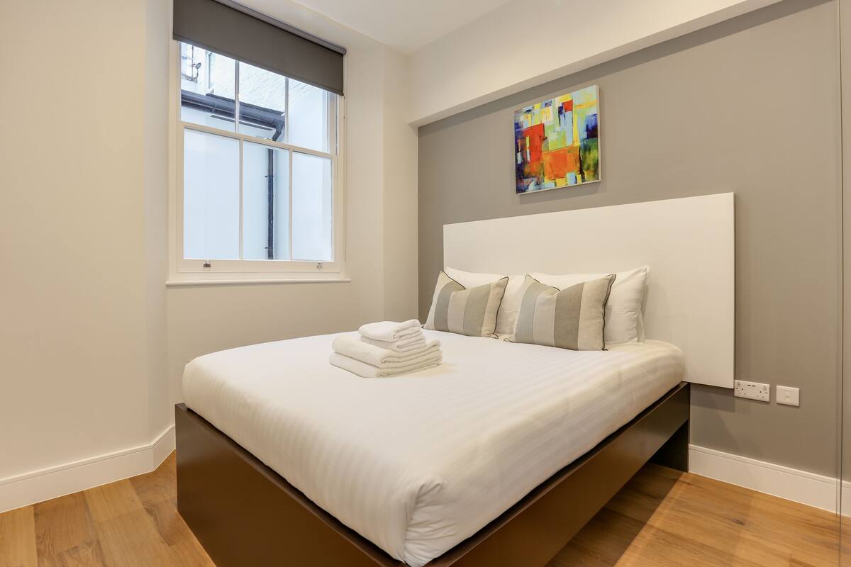 A modern bedroom with a large bed, a window with blinds, a colorful abstract painting on the wall, and a built-in bench under the bed.