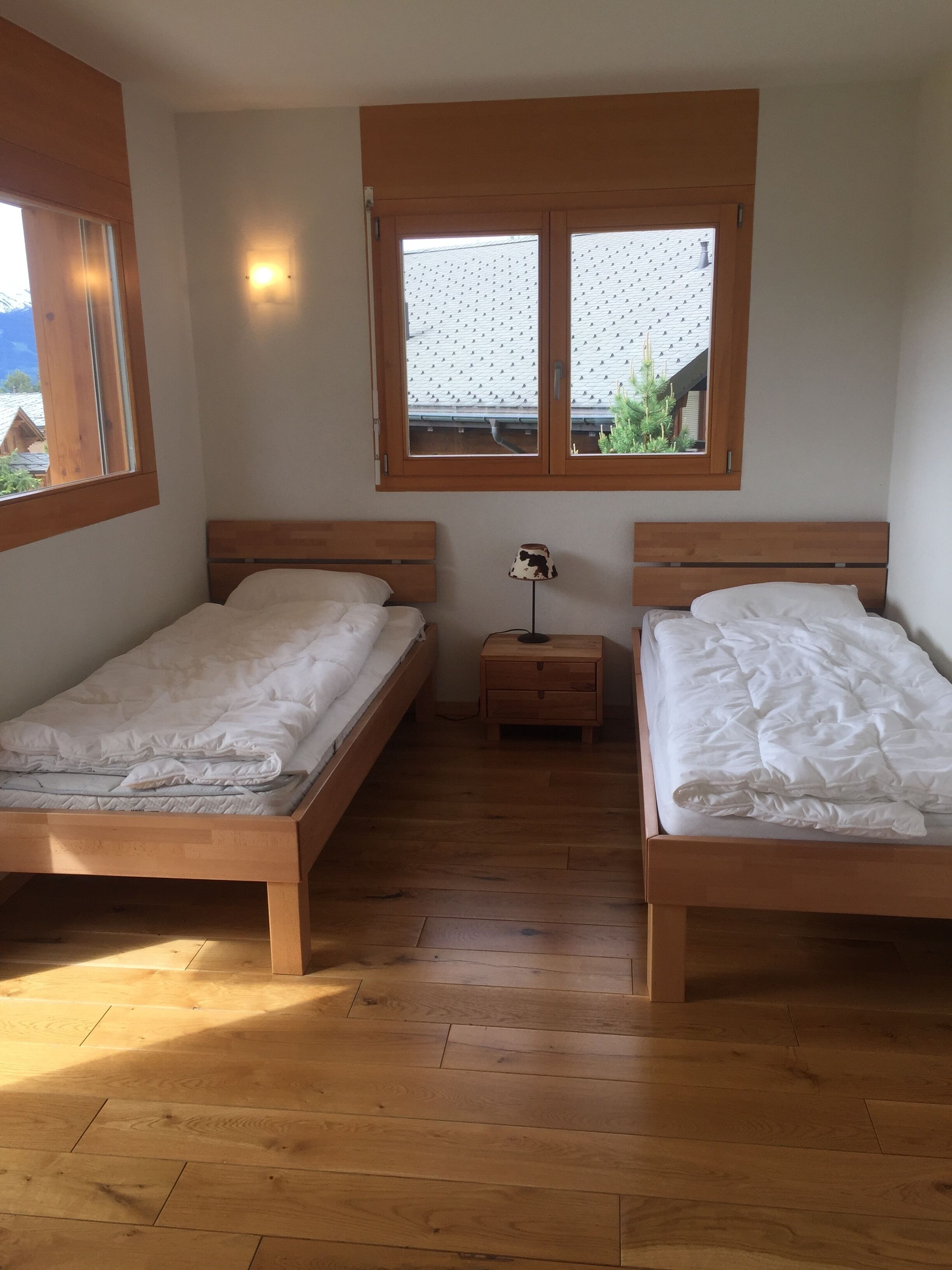 A bedroom with two wooden beds, a nightstand with a lamp, and a window with a view of a house and greenery.