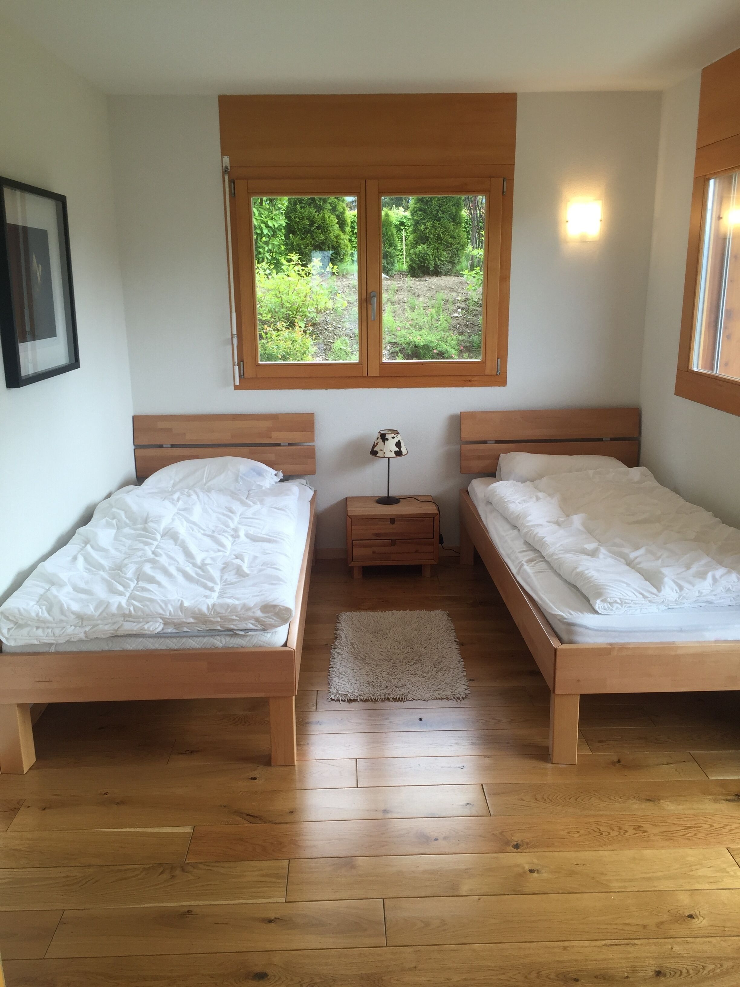 A room with two wooden beds, a small bedside table with a lamp, and a window with a view of greenery.