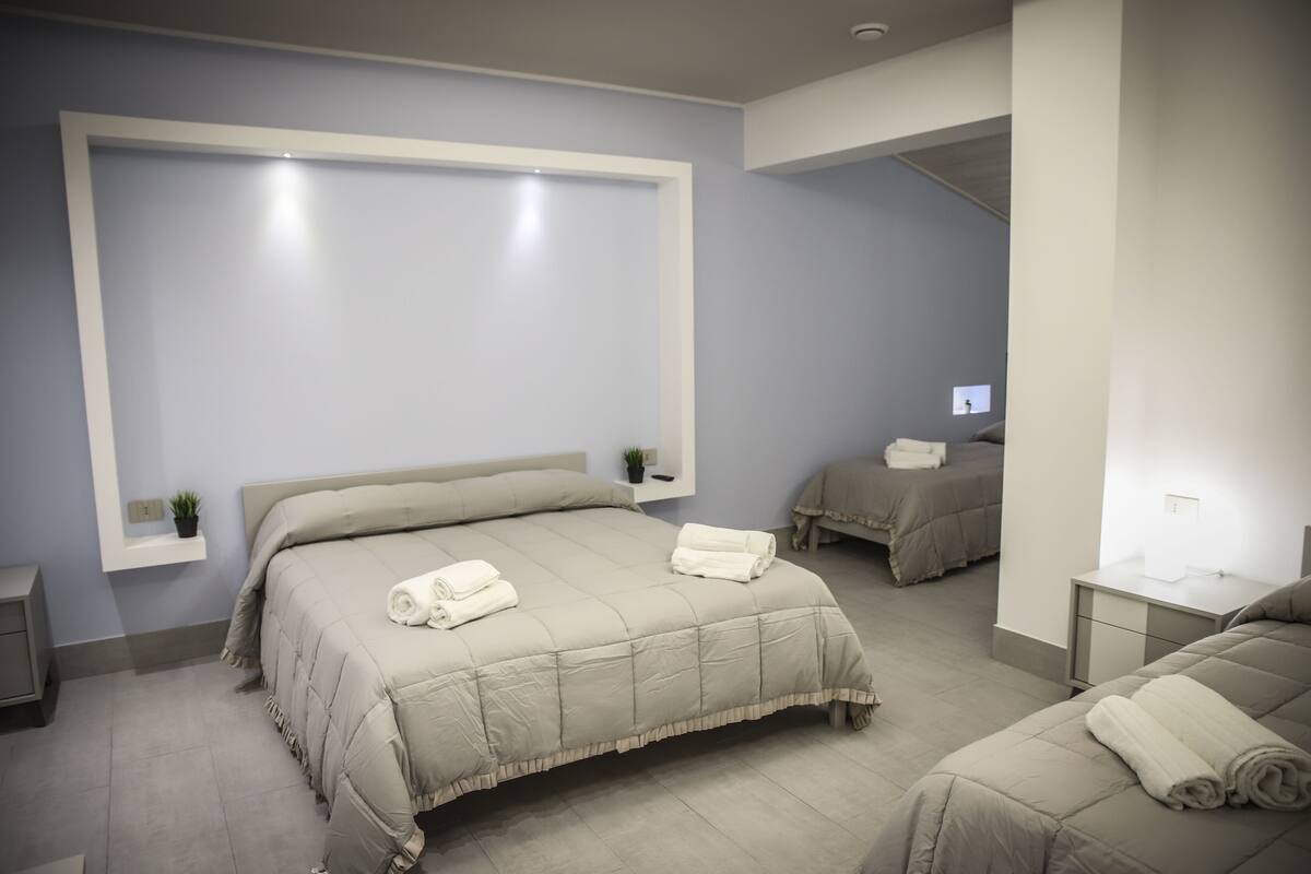 A modern hotel room with a large bed, a smaller bed, a bedside table, a nightstand, and a wall-mounted shelf with a plant.