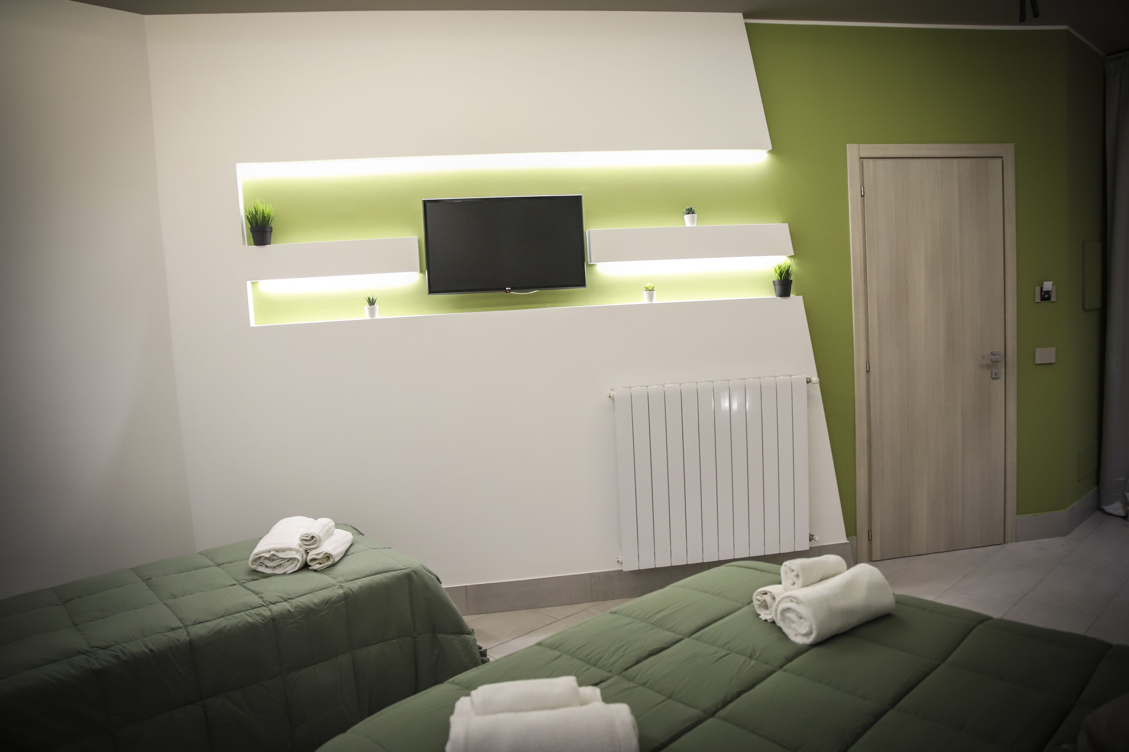 A hotel room with two green beds, a flat-screen TV mounted on a white wall, a radiator below, and a door to the right.