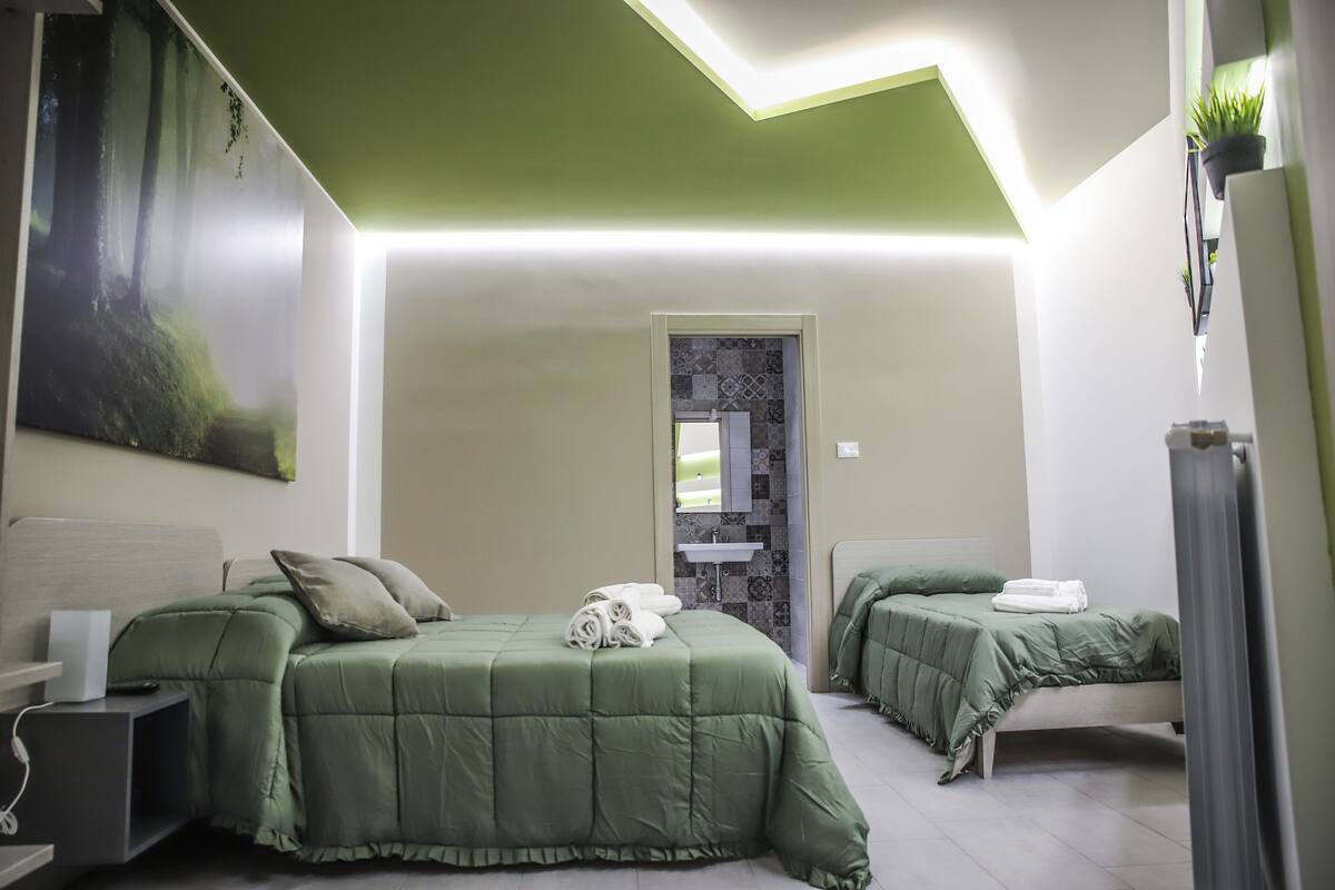 A modern hotel room with two beds, a large painting, a bathroom with a sink, and a green ceiling.