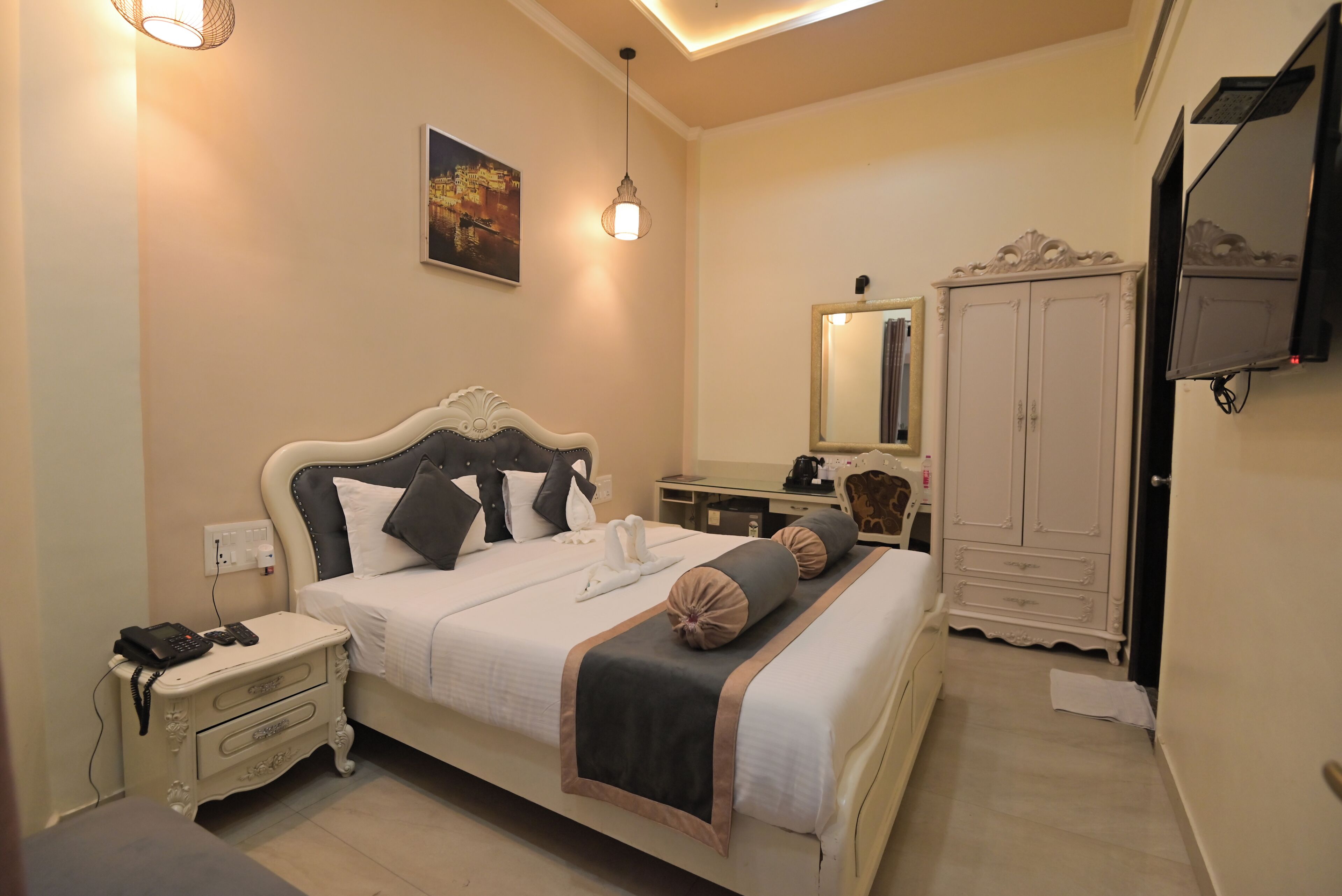 A hotel room with a bed, bedside table, lamp, wardrobe, and a painting on the wall.