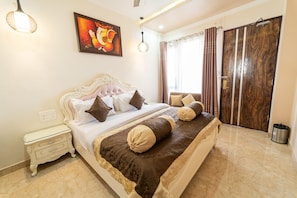 A bedroom with a large bed, a nightstand, a wall-mounted painting, and a wooden door. - Shree Ganesha Residency (Varanasi)