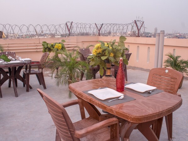 Un área de comedor en la azotea con una mesa dispuesta para dos personas, rodeada de sillas. Hay plantas en macetas y un jarrón decorativo sobre la mesa.