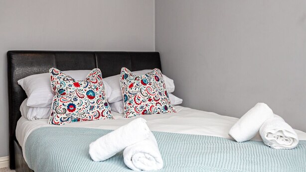 A neatly made bed with a blue blanket, two decorative pillows, and rolled white towels. - Irlam's Home from Home (Manchester)