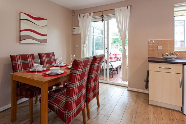 A dining area with a wooden table and plaid chairs, a kitchen with a tiled backsplash, and a sliding glass door leading to an outdoor area. - Irlam's Home from Home (Manchester)