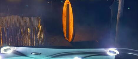 Un jacuzzi con una estructura de madera al fondo, un poste de madera y vistas a una orilla iluminada.