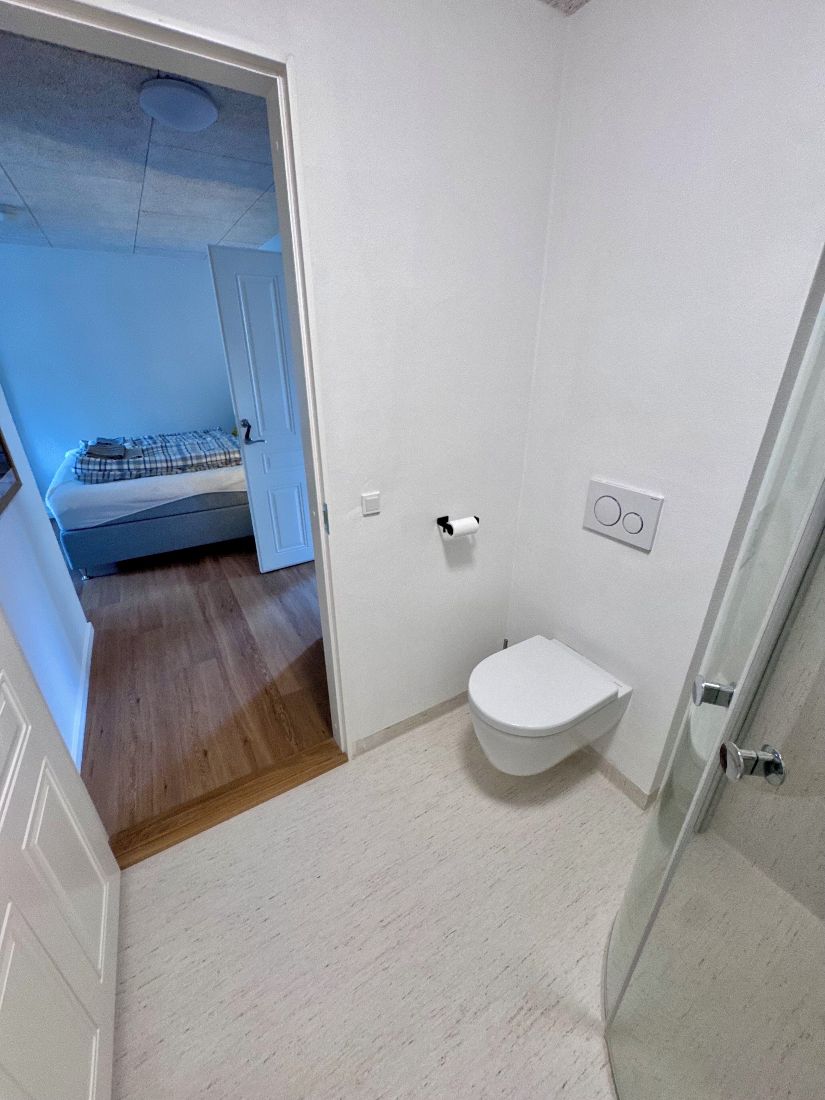 A bathroom with a toilet, a shower, and a door leading to a bedroom.
