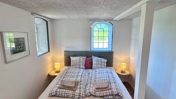Una cama bien hecha con cabecera, lámparas de noche, una ventana con vistas y un cuadro en la pared.