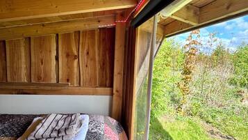 A wooden cabin interior with a bed, a window, and a view of greenery outside.