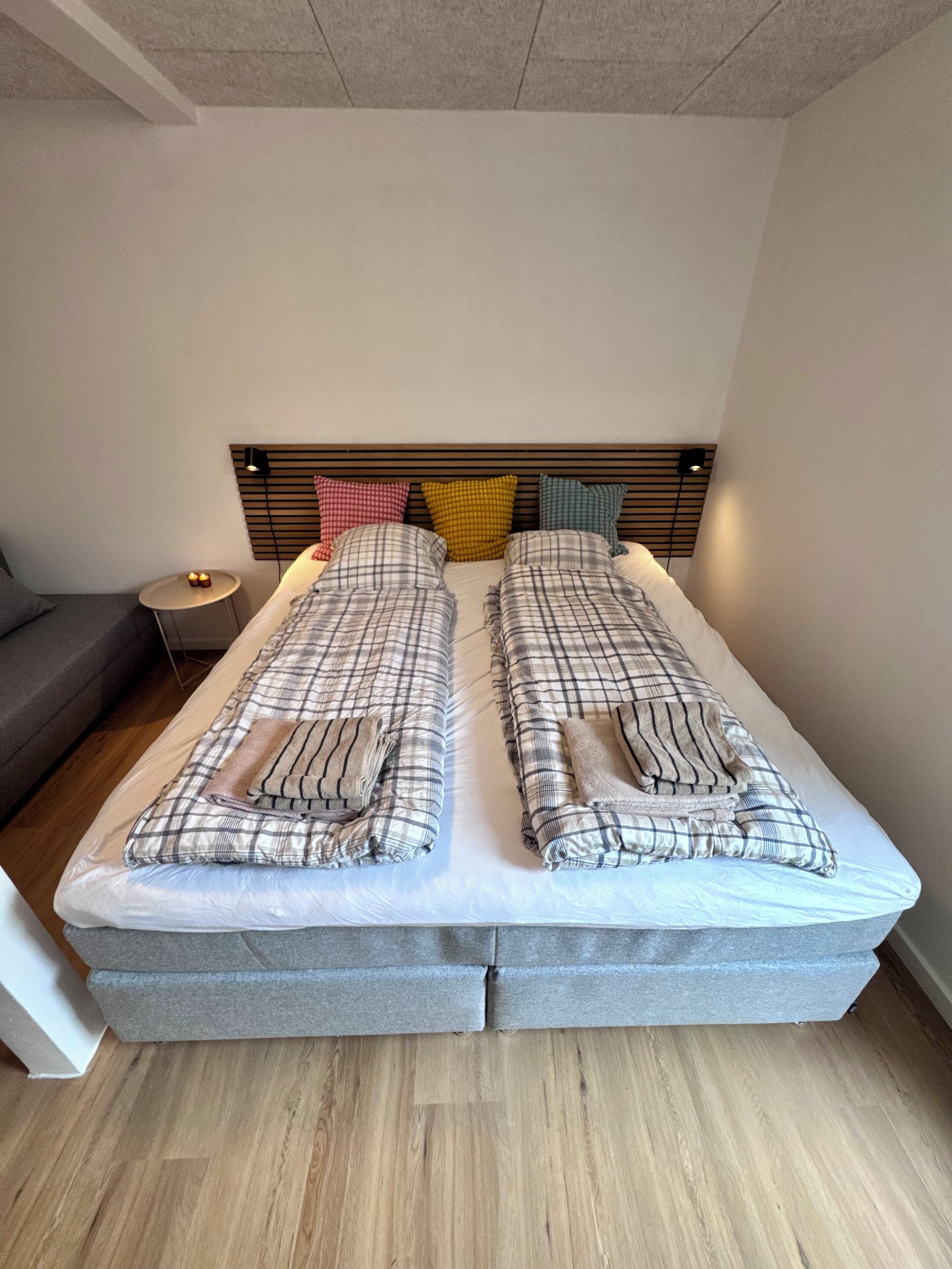 Una cama doble con cabecera, dos almohadas, una mesita de noche con lámpara y un sofá gris.