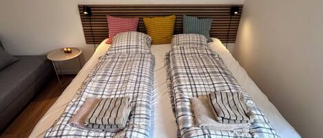 Una cama doble con cabecera, dos almohadas, una mesita de noche con lámpara y un sofá gris.