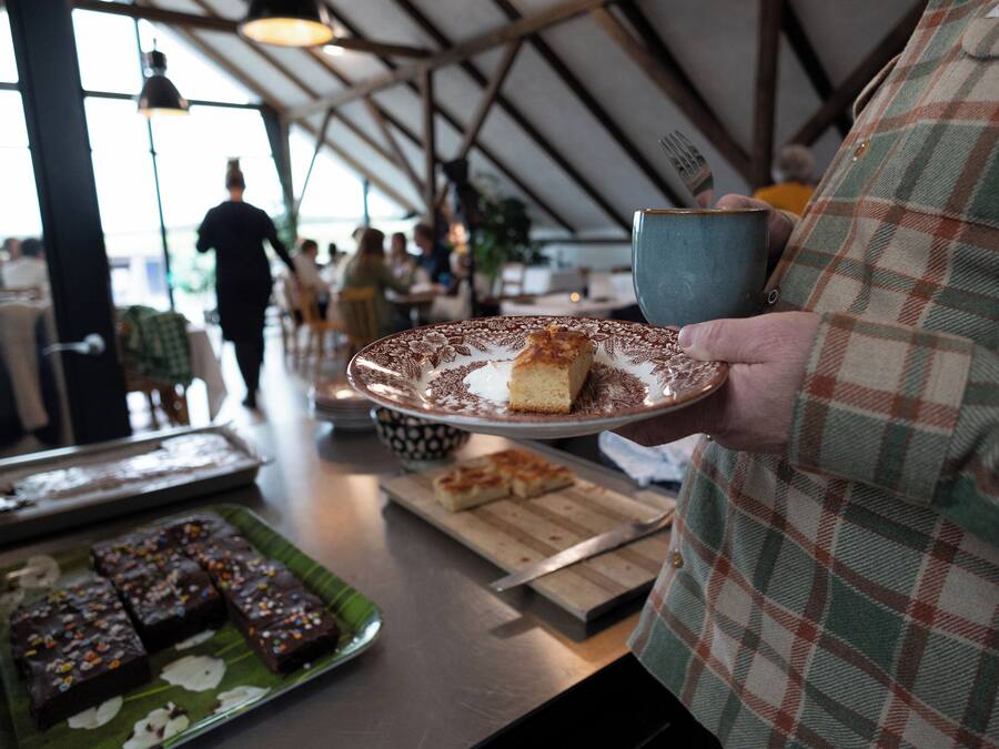 Una persona sosteniendo un plato con un postre y una taza, con una bandeja de brownies y una tabla de cortar con un cuchillo sobre una mesa en un restaurante.