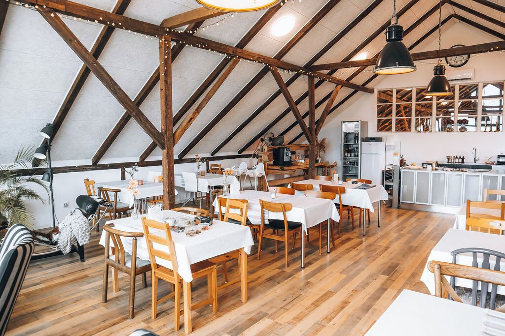 A spacious dining area with wooden tables and chairs, a kitchen in the background, and a high ceiling with exposed beams.