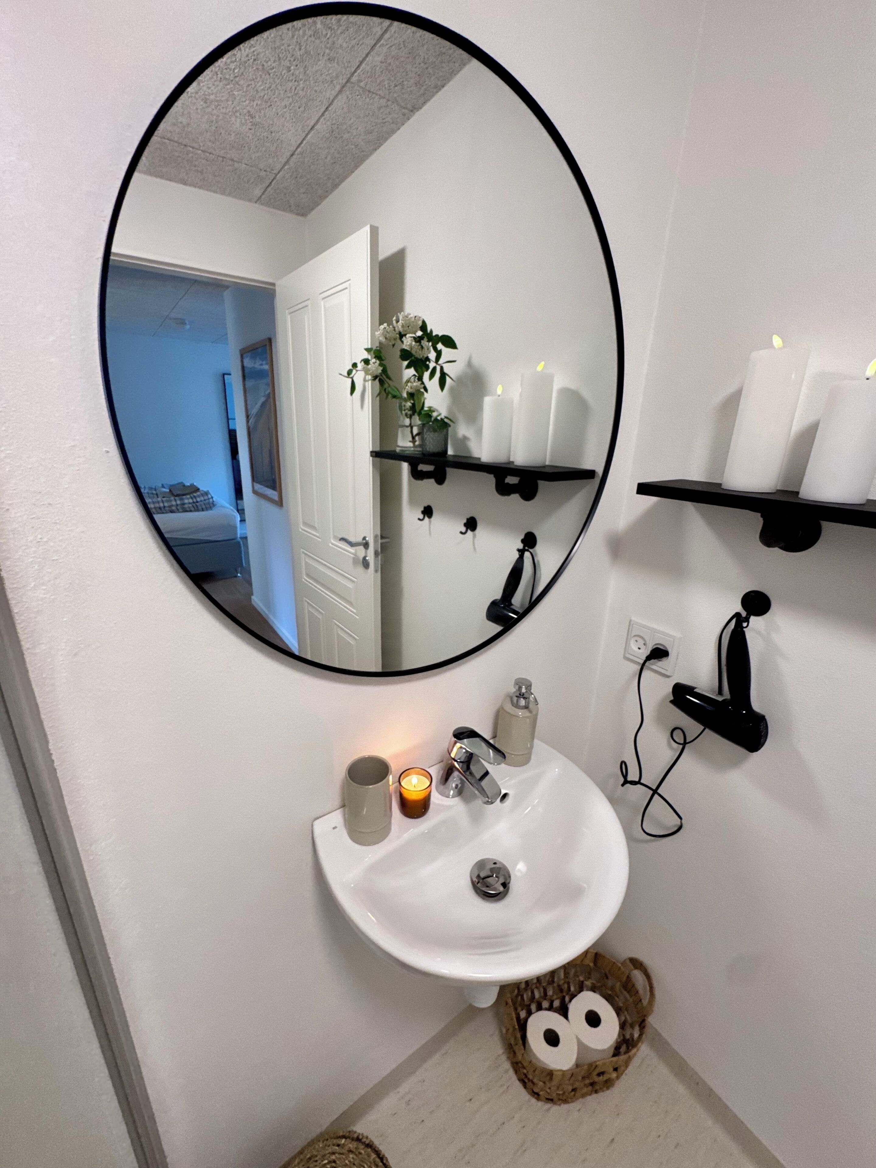 Une salle de bain avec un miroir rond, un lavabo blanc et deux étagères sur lesquelles sont posées des bougies.