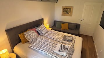 A bedroom with a bed, a bedside table with a lamp, a sofa, a door, and a framed picture on the wall.