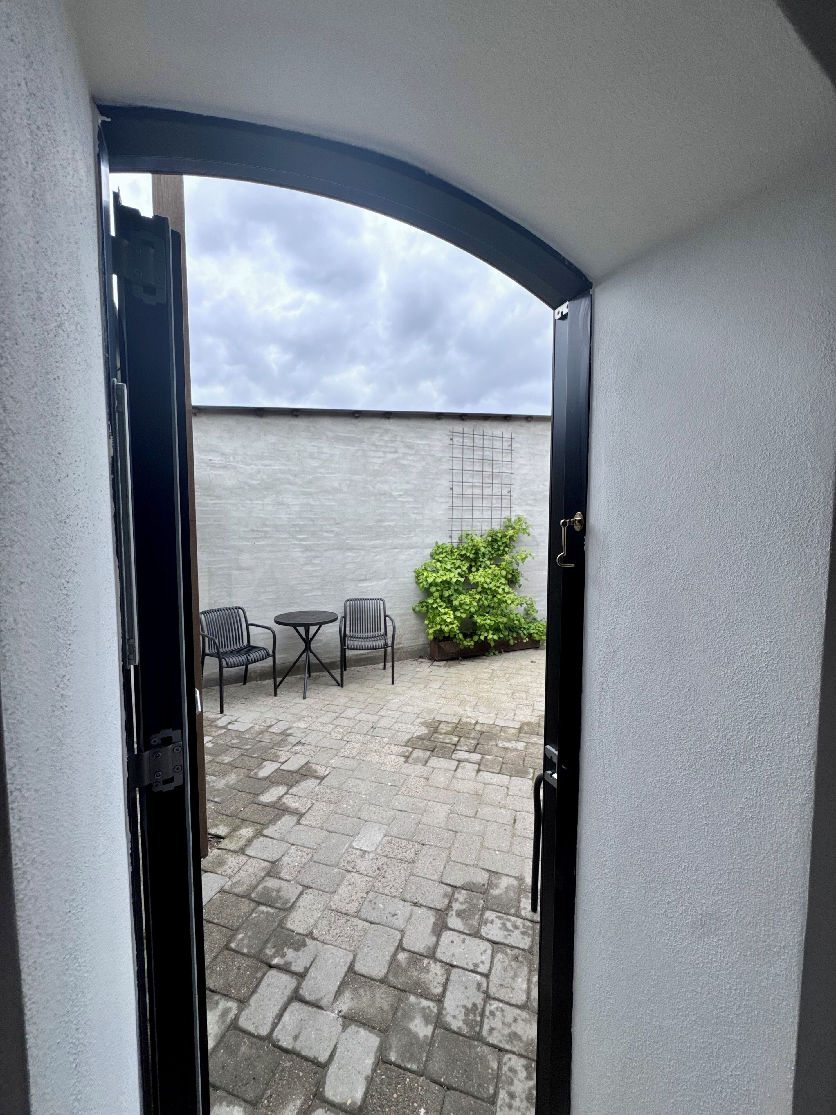 An open door to a courtyard with a white wall, two chairs, a small table, and a potted plant.