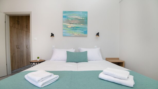 Un lit bien fait, avec du linge de lit blanc et un plaid turquoise, une petite table de chevet avec une lampe et une armoire en bois.