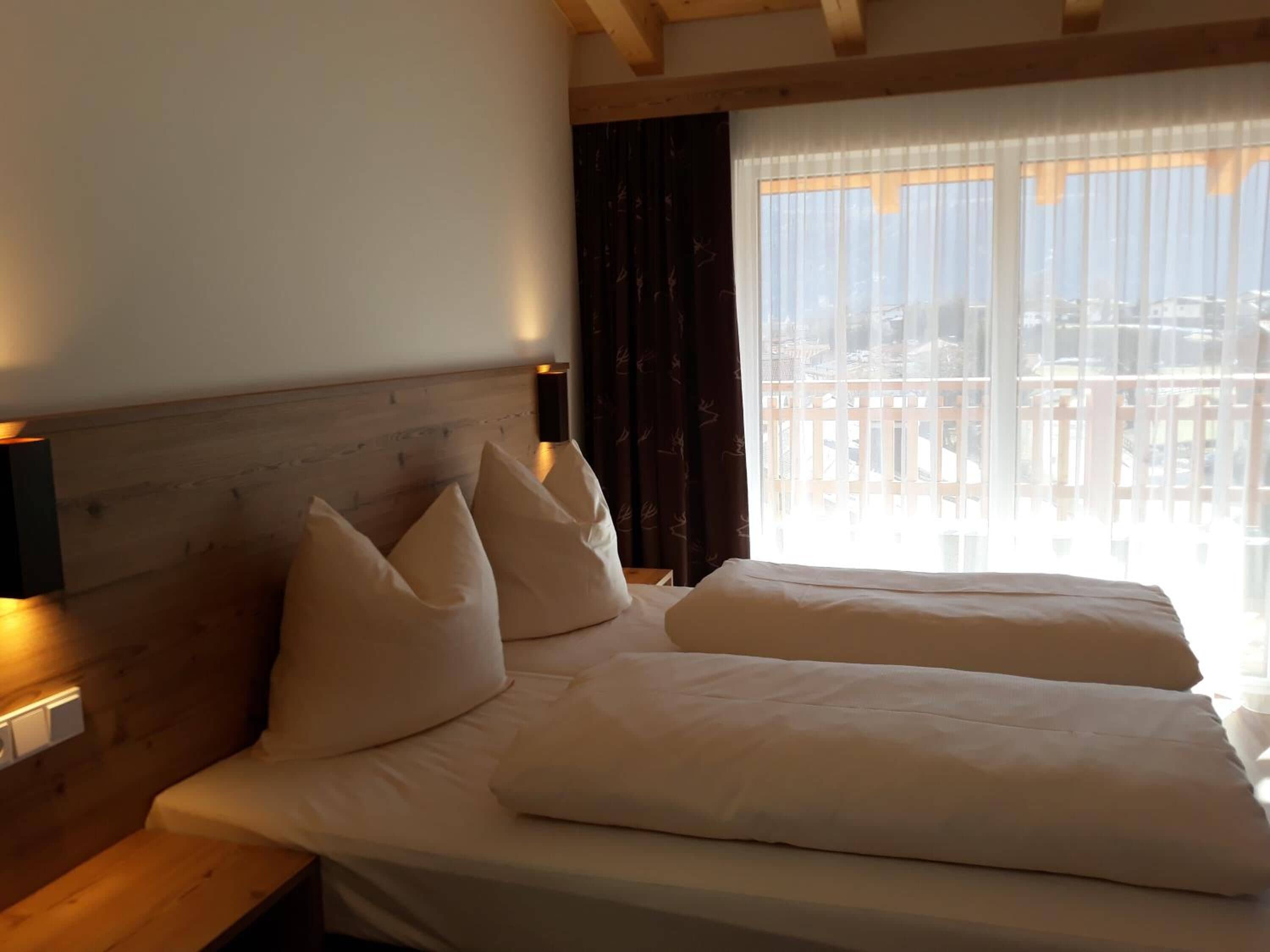 Une chambre d’hôtel avec une tête de lit en bois, deux lits avec du linge de lit blanc, une fenêtre avec des rideaux légers et une vue sur les bâtiments environnants.