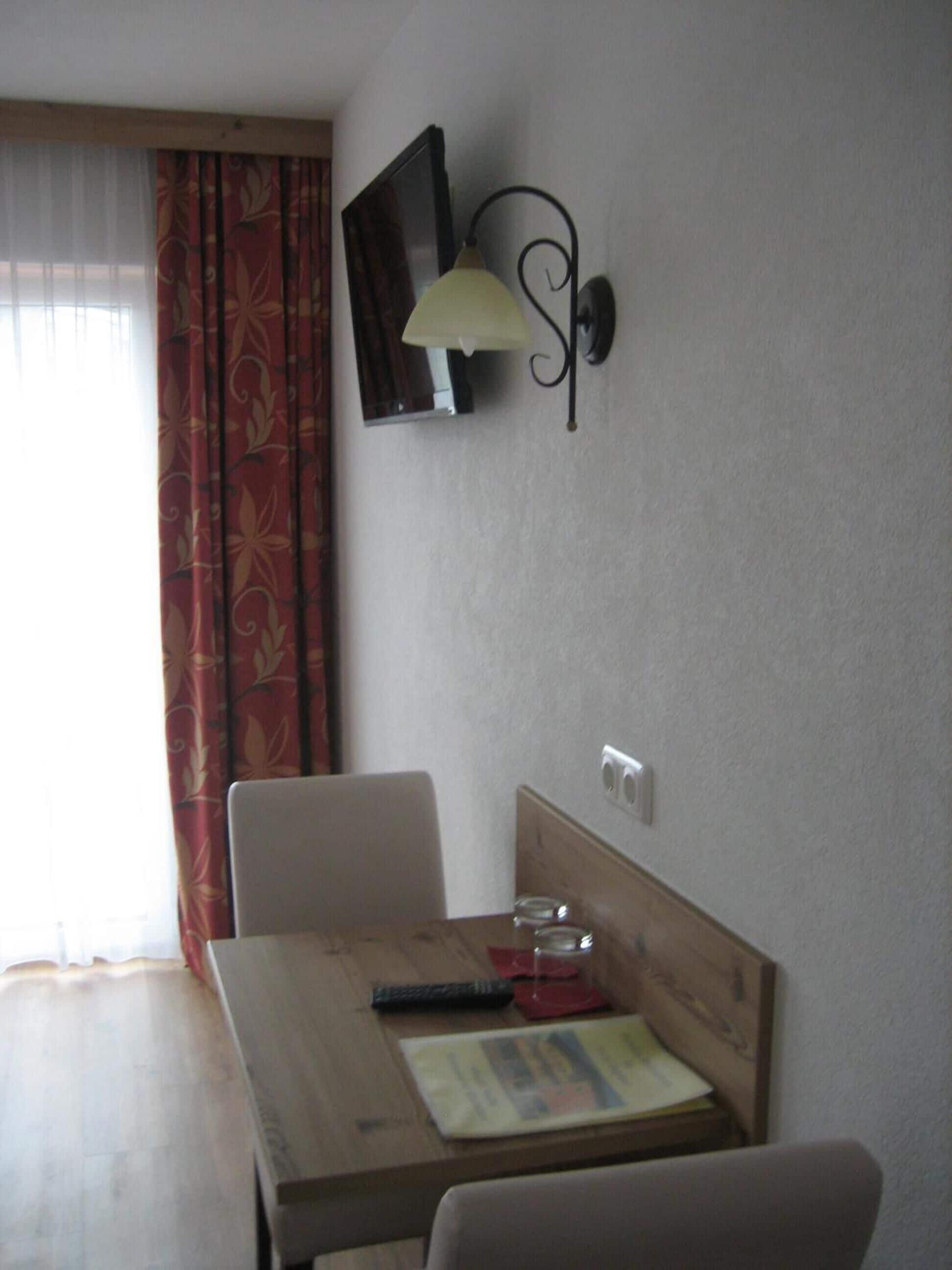 A small dining area with a wooden table, two chairs, a wall-mounted TV, and a wall lamp.