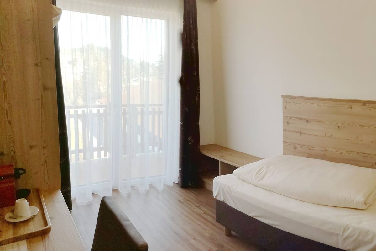 A bedroom with a wooden headboard, a bed with white bedding, a bench, and a window with sheer curtains.