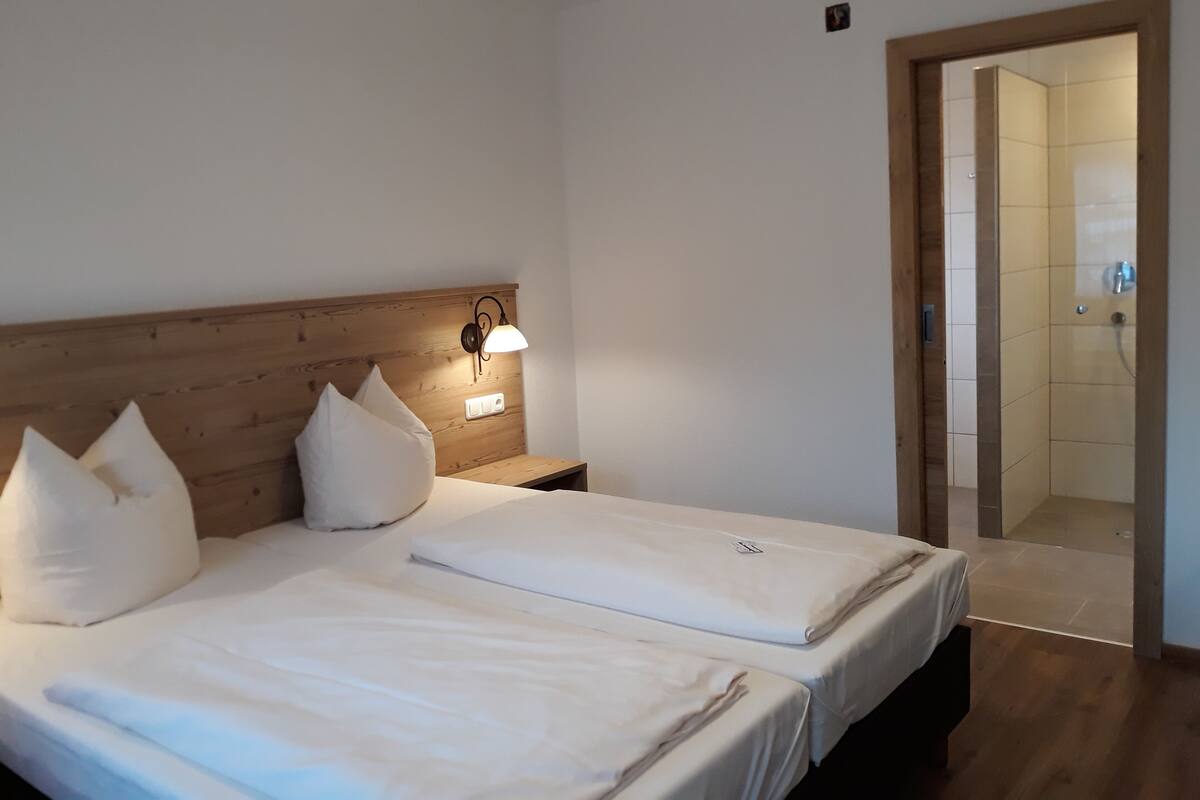 A hotel room with two beds, a wooden headboard, a nightstand with a lamp, and a bathroom visible through an open door.
