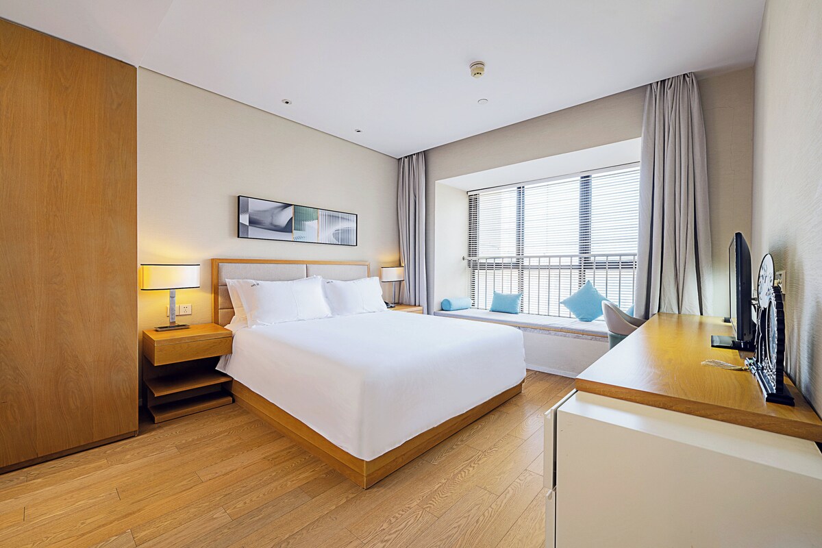 A hotel room with a large bed, bedside tables, a desk, a window with curtains, and a mounted television.