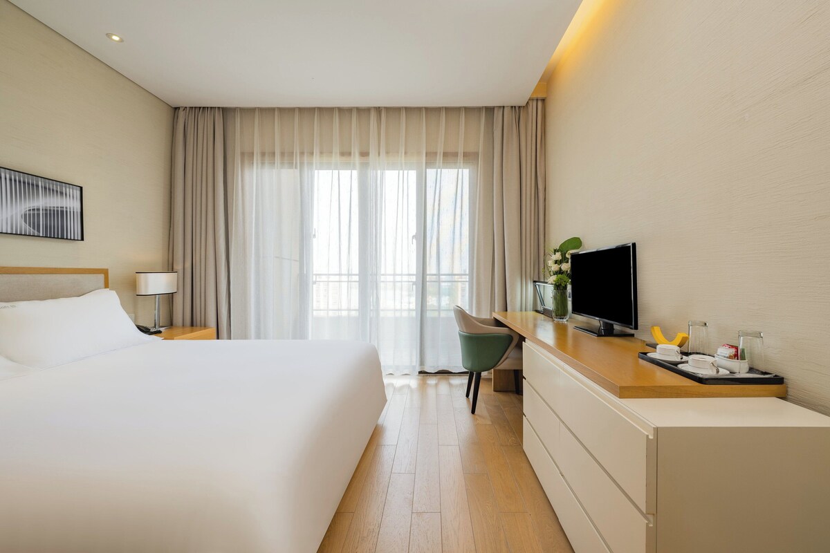 A hotel room with a large bed, a desk with a TV, a chair, and a window with curtains.