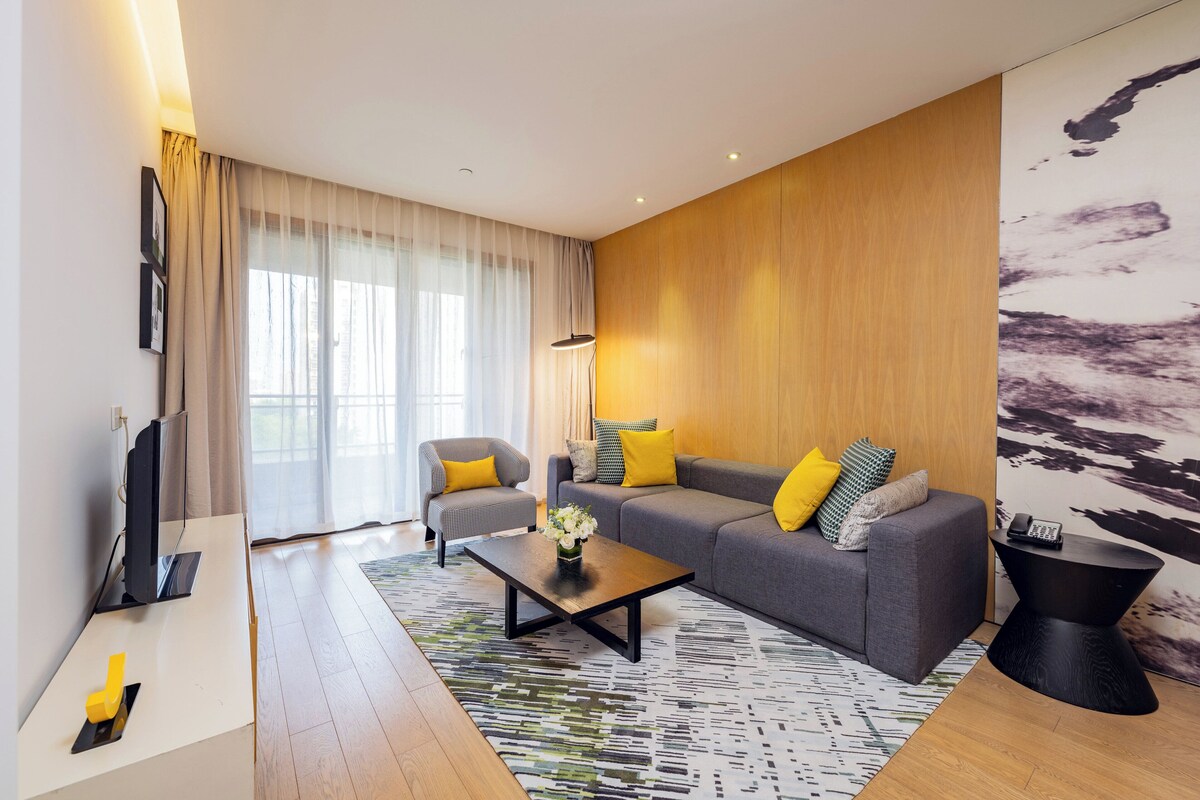 A modern living room with a grey sofa, two yellow pillows, a coffee table, a TV, and a large window with sheer curtains.