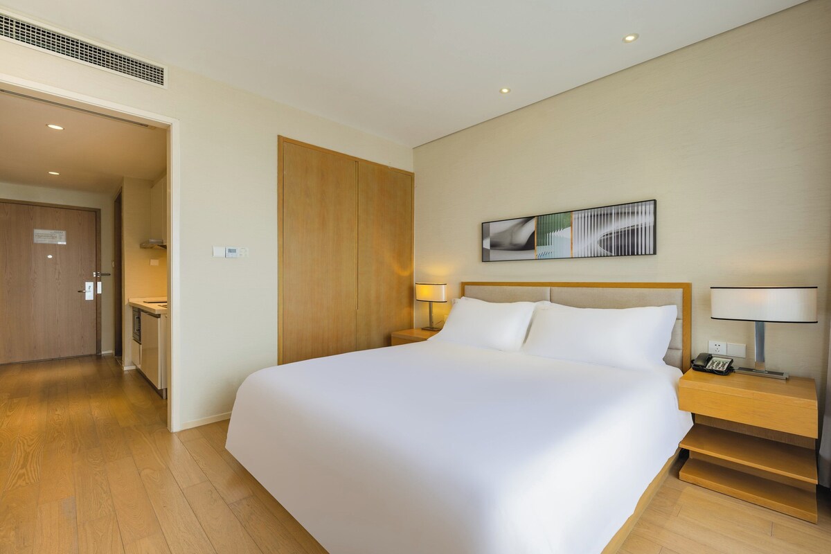 A hotel room with a large bed, a bedside table with a lamp, a small desk with a chair, and a wooden wardrobe.