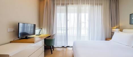 Una habitación de hotel moderna con una cama, un escritorio con computadora, un televisor y una ventana con cortinas translúcidas.