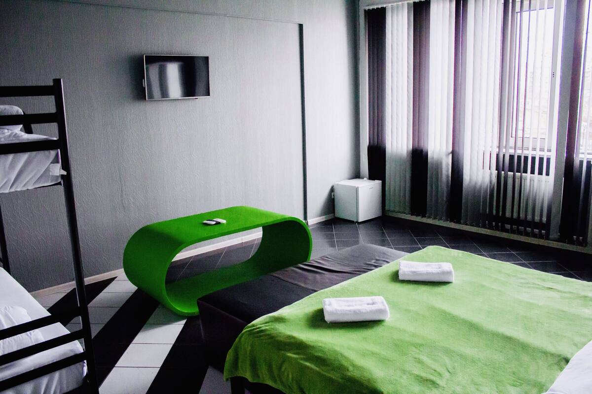 A hotel room with a bunk bed, a green ottoman, a flat-screen TV, a radiator, and a window with curtains.