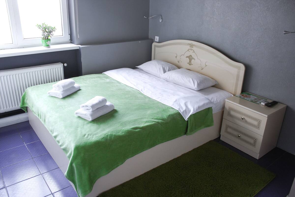 A neatly made bed with green and white bedding, a bedside table with a book, and a window with a plant.