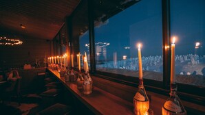 A bar with candles and bottles, a view of a snowy landscape through large windows, and a wooden interior. - Little Peak (Kuusamo)