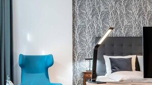 A hotel room with a modern bed, a blue armchair, a small table, and a nightstand with a lamp. - H23 Boardinghotel (Stuttgart)