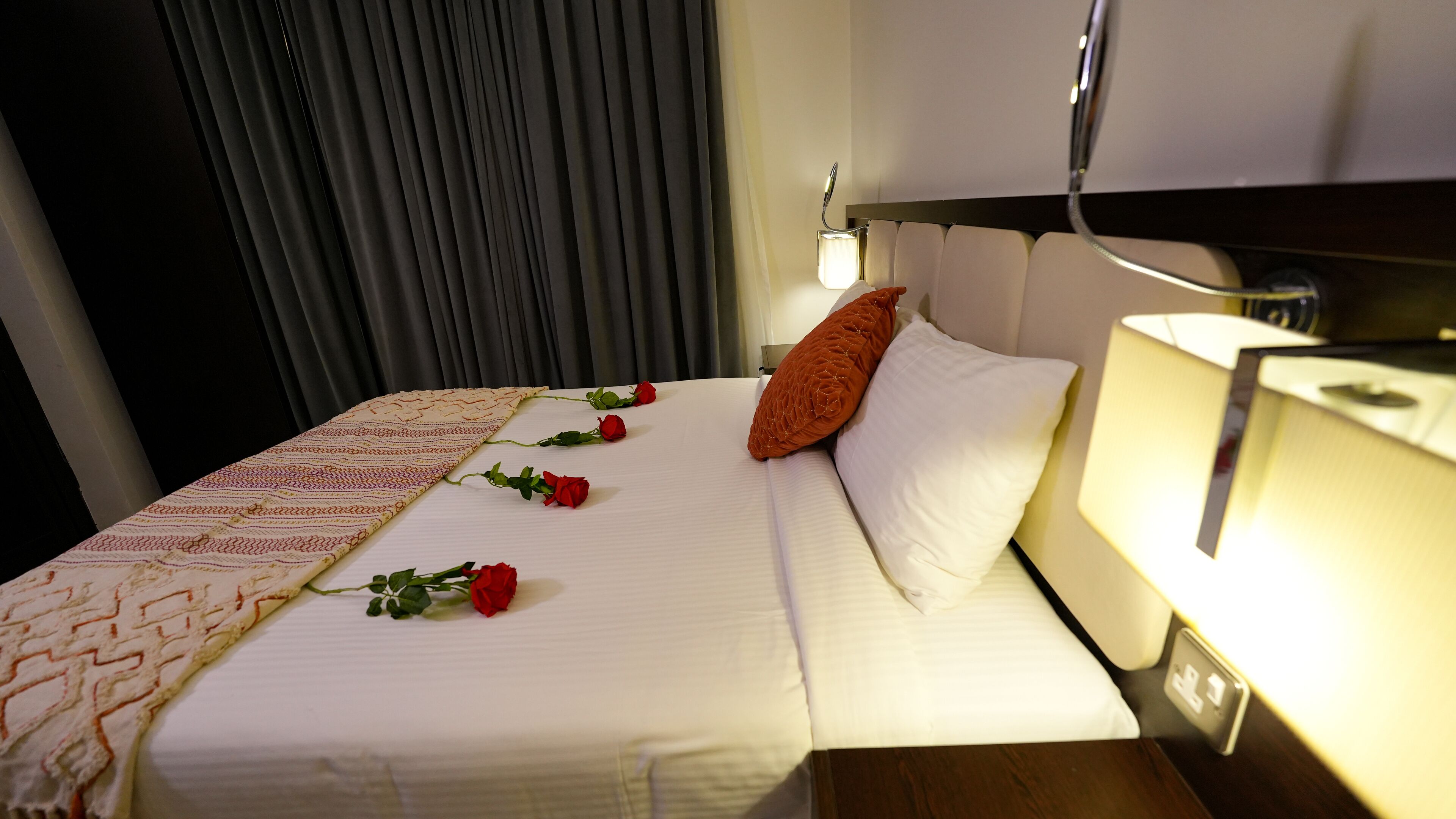 Un lit bien fait, recouvert d’une couvre-lit à motifs, avec un oreiller blanc et deux roses rouges.