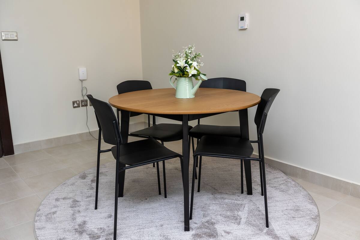 Uma mesa de madeira redonda com um vaso de flores brancas, rodeada por quatro cadeiras pretas sobre um tapete cinzento.